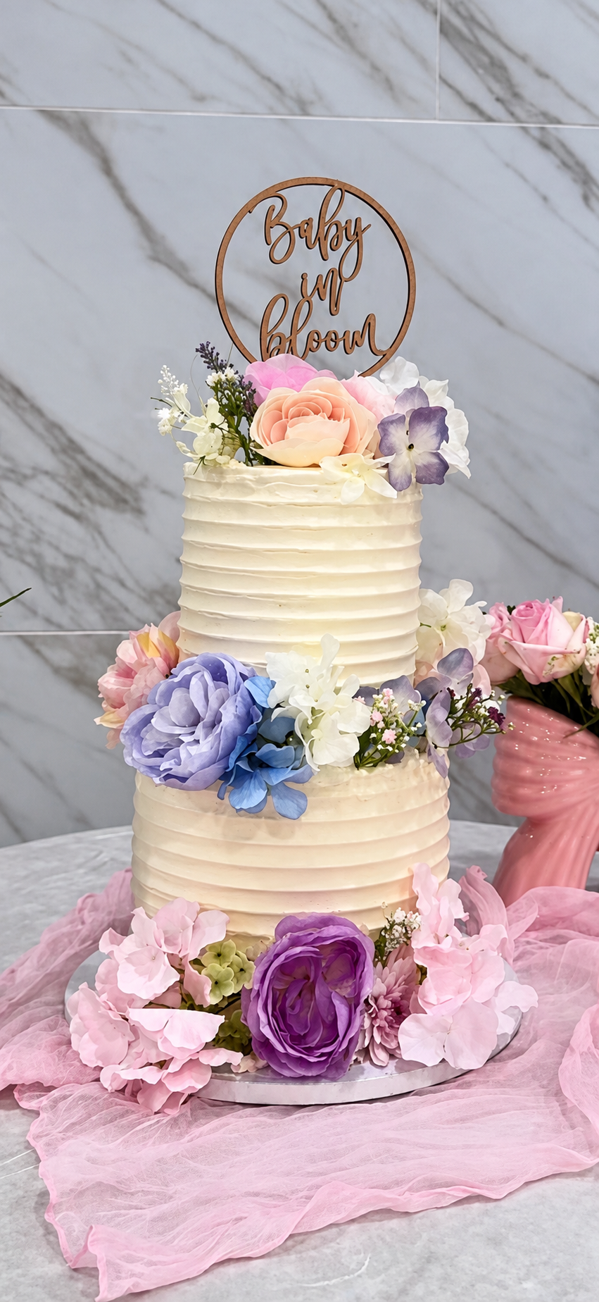 LovelyEatz two-tier ivory cake with pink, blue, and purple flowers and a Baby in bloom topper